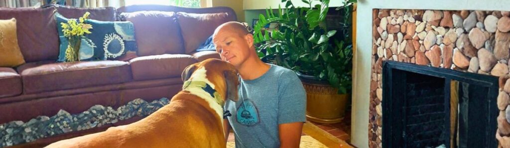 Holistic veterinarian sitting on a living room rug with a large tan and white dog, sharing a calm moment beside a stone fireplace